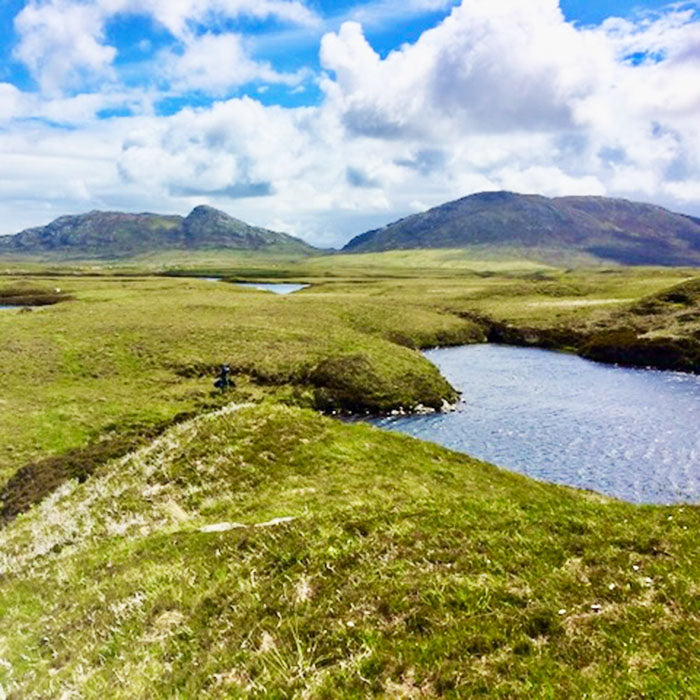 A scenic loch landscape photograph of a grassy valley with a winding blue river, distant mountains, and a bright blue sky filled with white clouds.